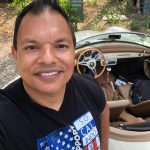 Man smiling in front of a vintage car with a beige interior, outdoors with trees and a sign in the background.