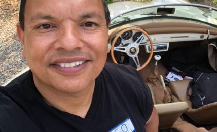 Man smiling in front of a vintage car with a beige interior, outdoors with trees and a sign in the background.