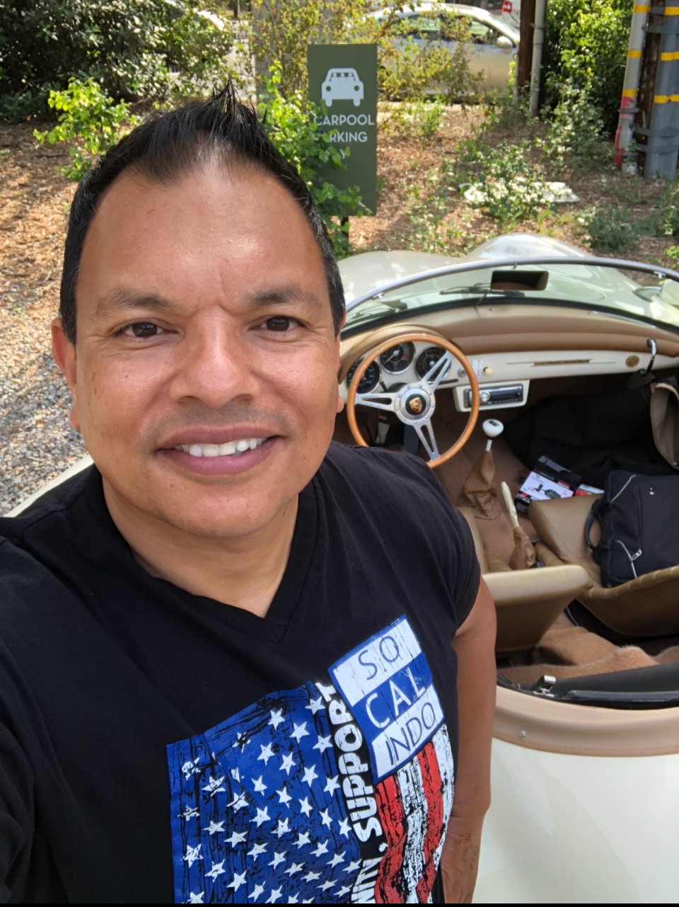 Man smiling in front of a vintage car with a beige interior, outdoors with trees and a sign in the background.