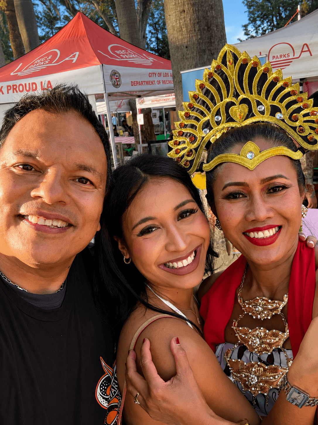 Three people smiling outdoors, one wearing a traditional headdress, with tents and trees in the background.