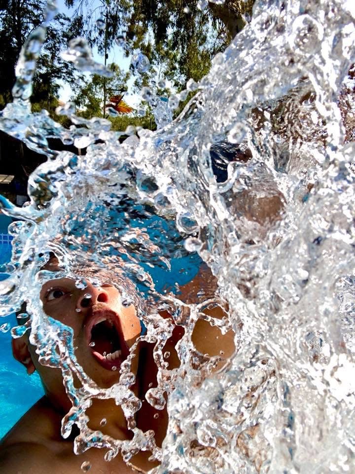 Child with mouth open, splashing water outdoors with trees and sky visible in background.