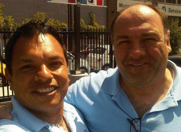 Two men smiling outdoors, one with dark hair and the other bald, both wearing light blue shirts.