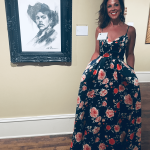 Woman in a floral dress standing next to a framed black and white portrait in an art gallery.