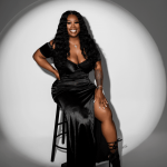 Woman sitting on a stool, smiling, wearing a black dress and high-heeled boots, in a circular spotlight.