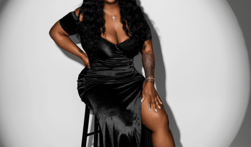 Woman sitting on a stool, smiling, wearing a black dress and high-heeled boots, in a circular spotlight.
