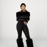 Woman in black outfit with fur accents on arms and legs, standing against a plain background.