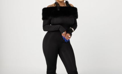 Woman in black outfit with fur accents on arms and legs, standing against a plain background.
