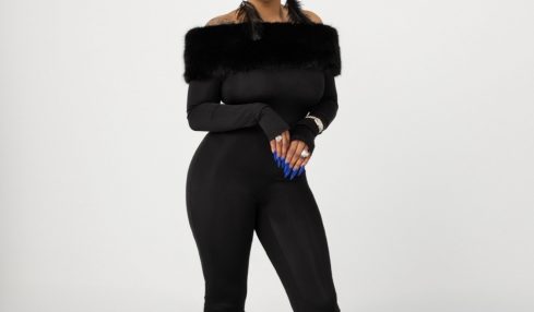Woman in black outfit with fur accents on arms and legs, standing against a plain background.