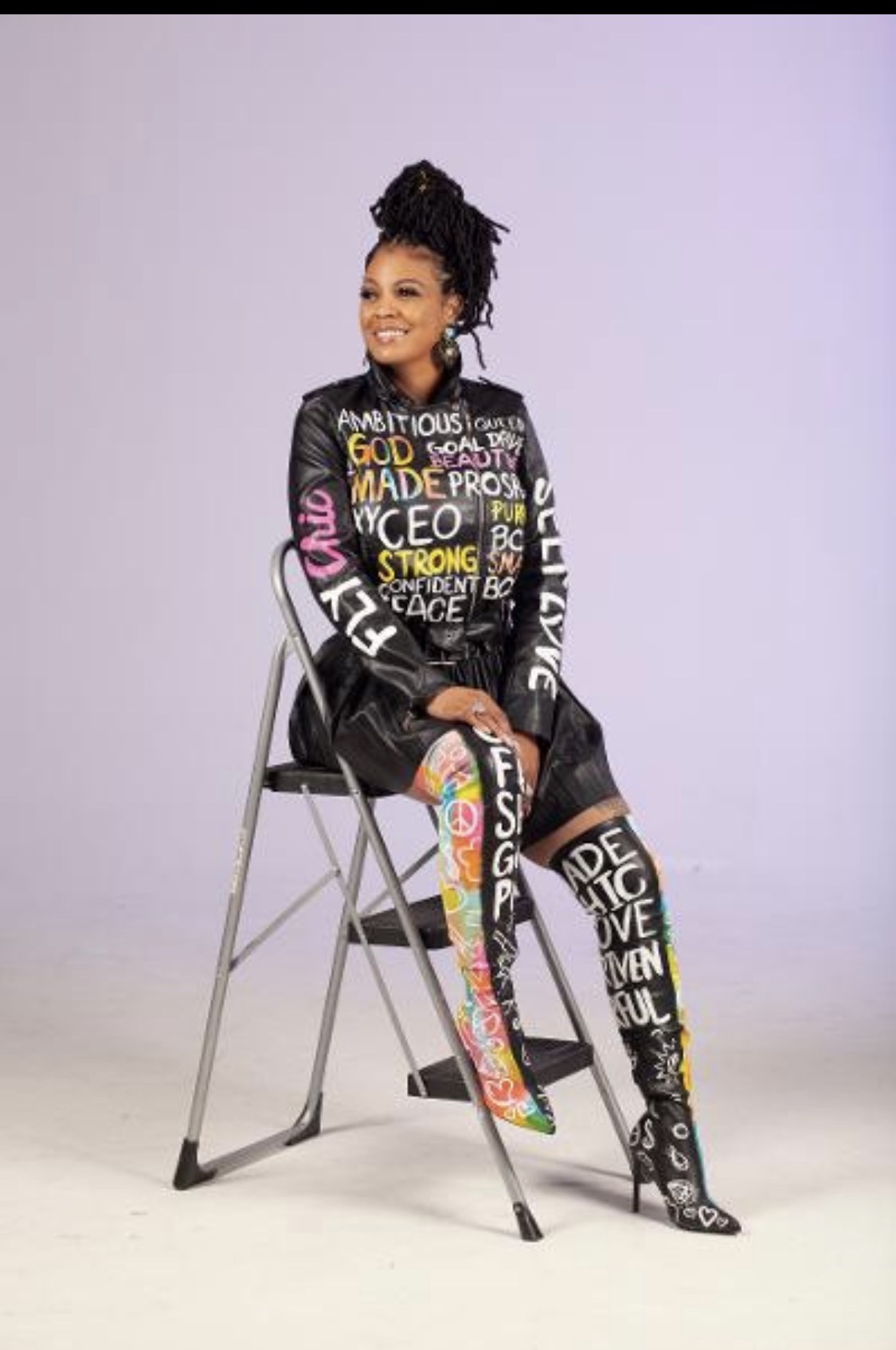 Woman sitting on a chair, smiling, wearing a colorful, text-covered outfit, against a plain background.