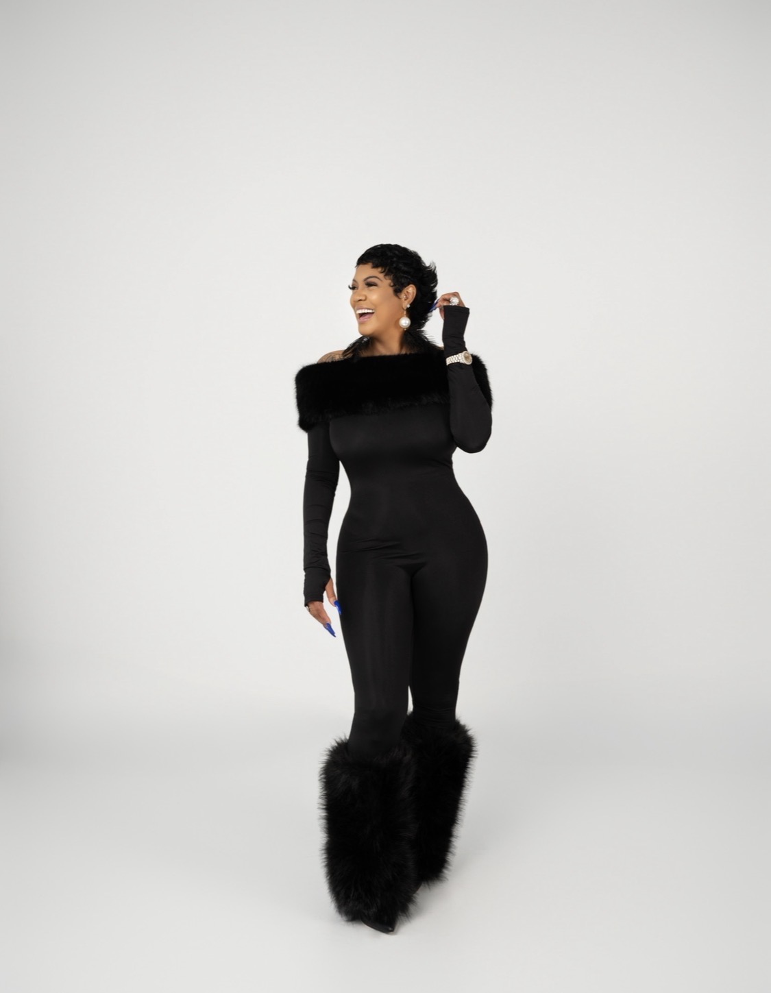 Woman in black outfit with furry boots smiling and holding a phone, standing against a plain background.