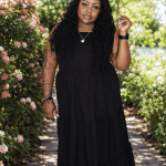 Woman in black dress walking on a garden path with pink flowers and green trees, smiling, holding a bracelet.