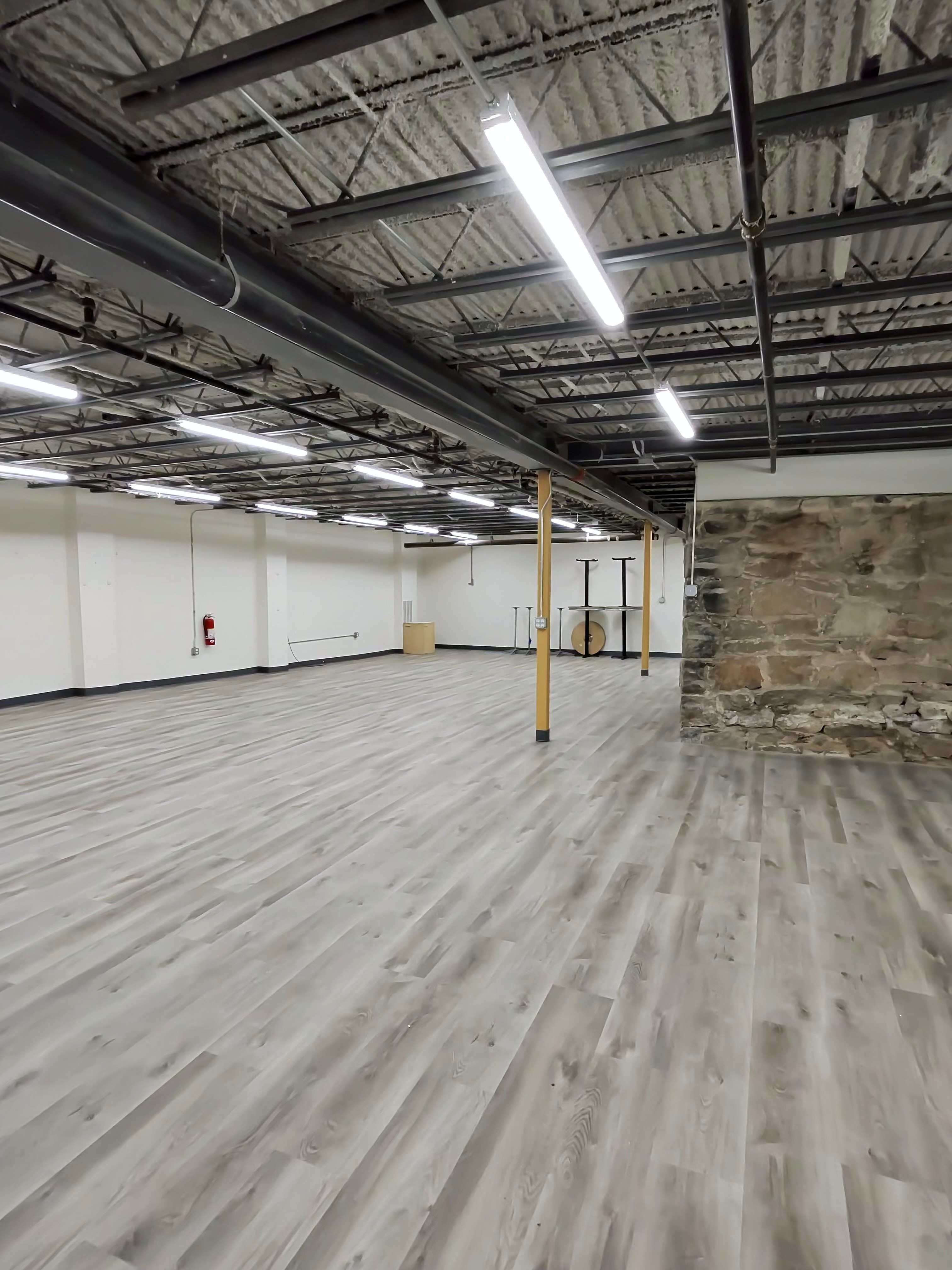 Empty industrial space with high ceiling, fluorescent lights, wooden floor, and a brick wall on the right.