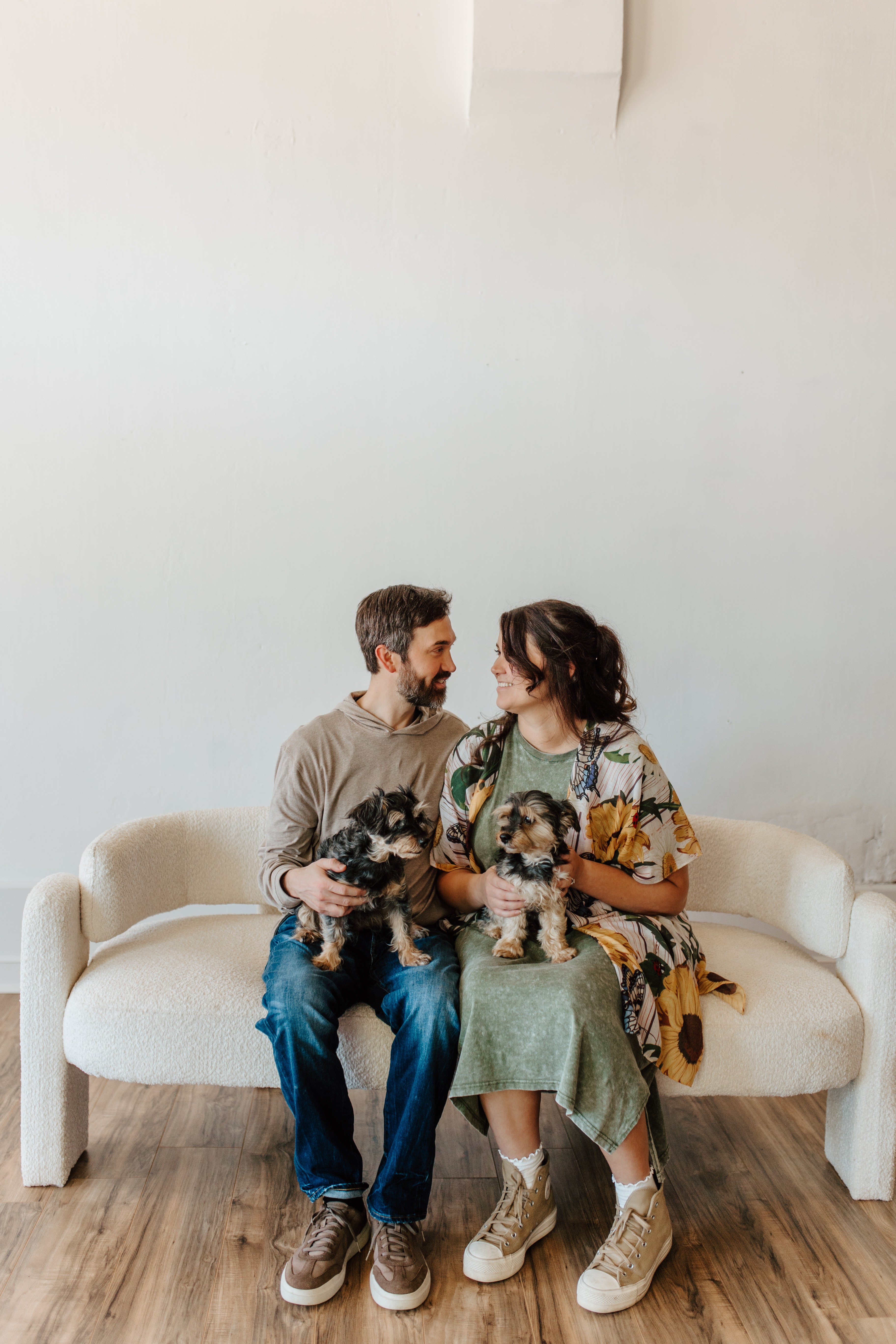 A couple sitting on a white sofa holding two small dogs, with a plain white wall background.