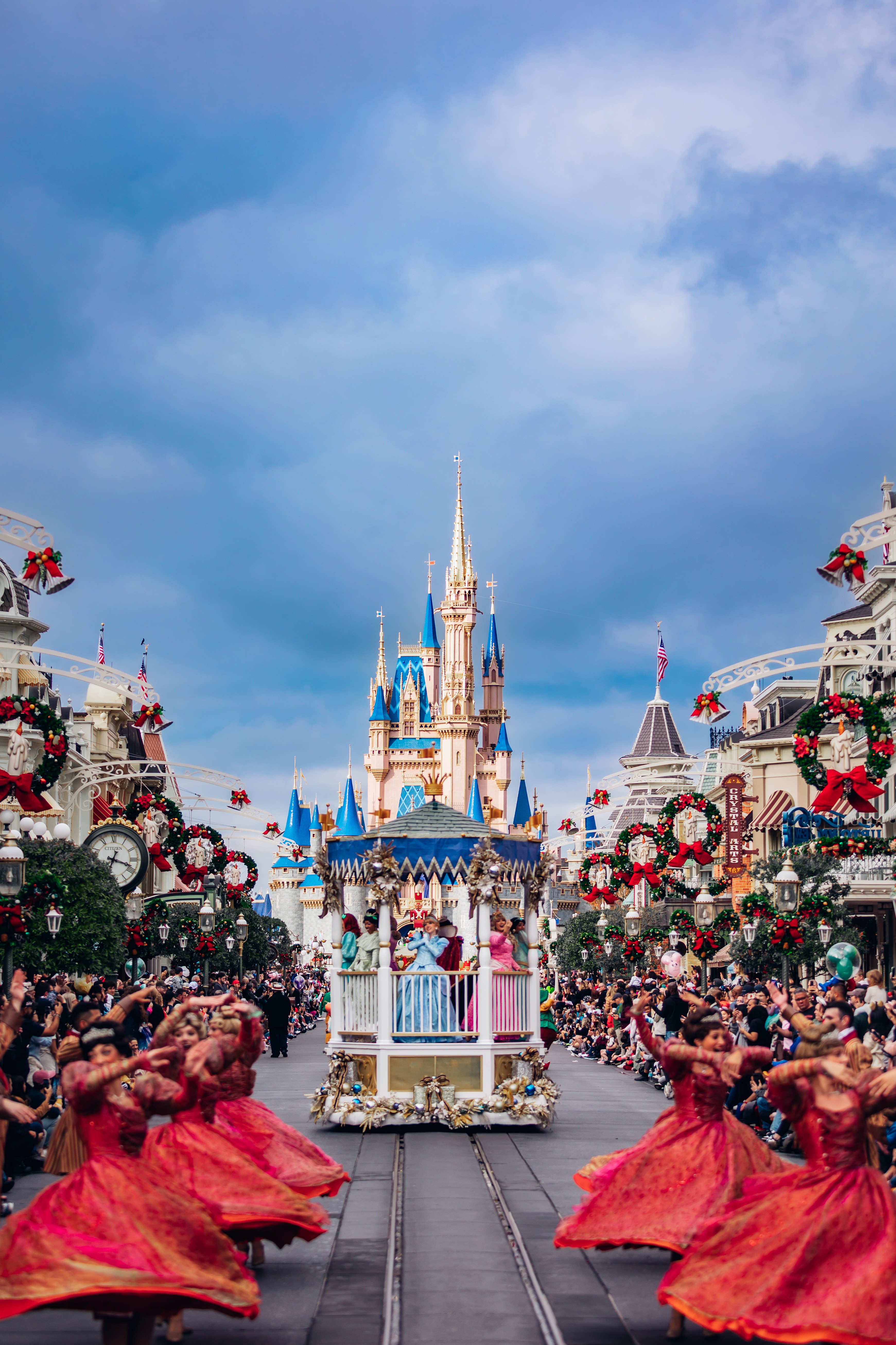 Castle with blue spires and parade with dancers in red dresses, decorated with wreaths and flowers, under cloudy sky.