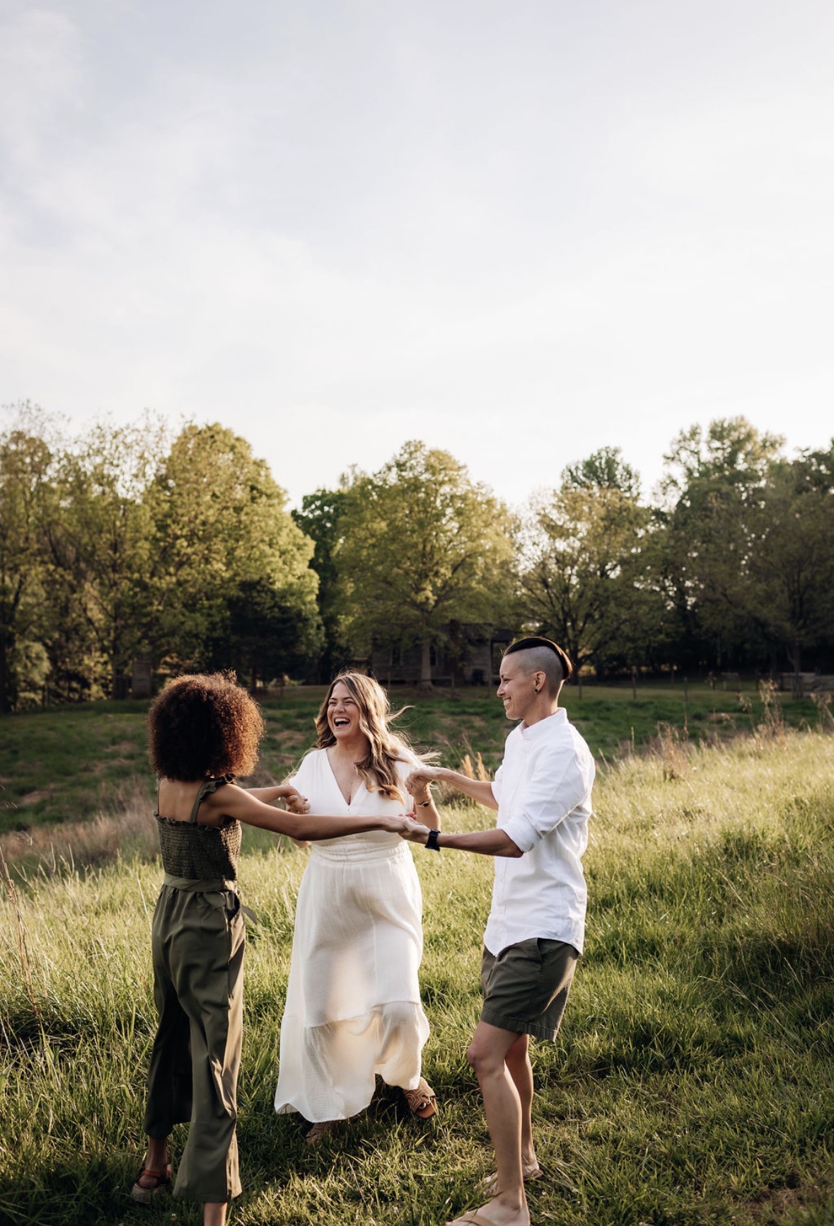 Three people holding hands and dancing in a grassy field with trees and a cloudy sky in the background.