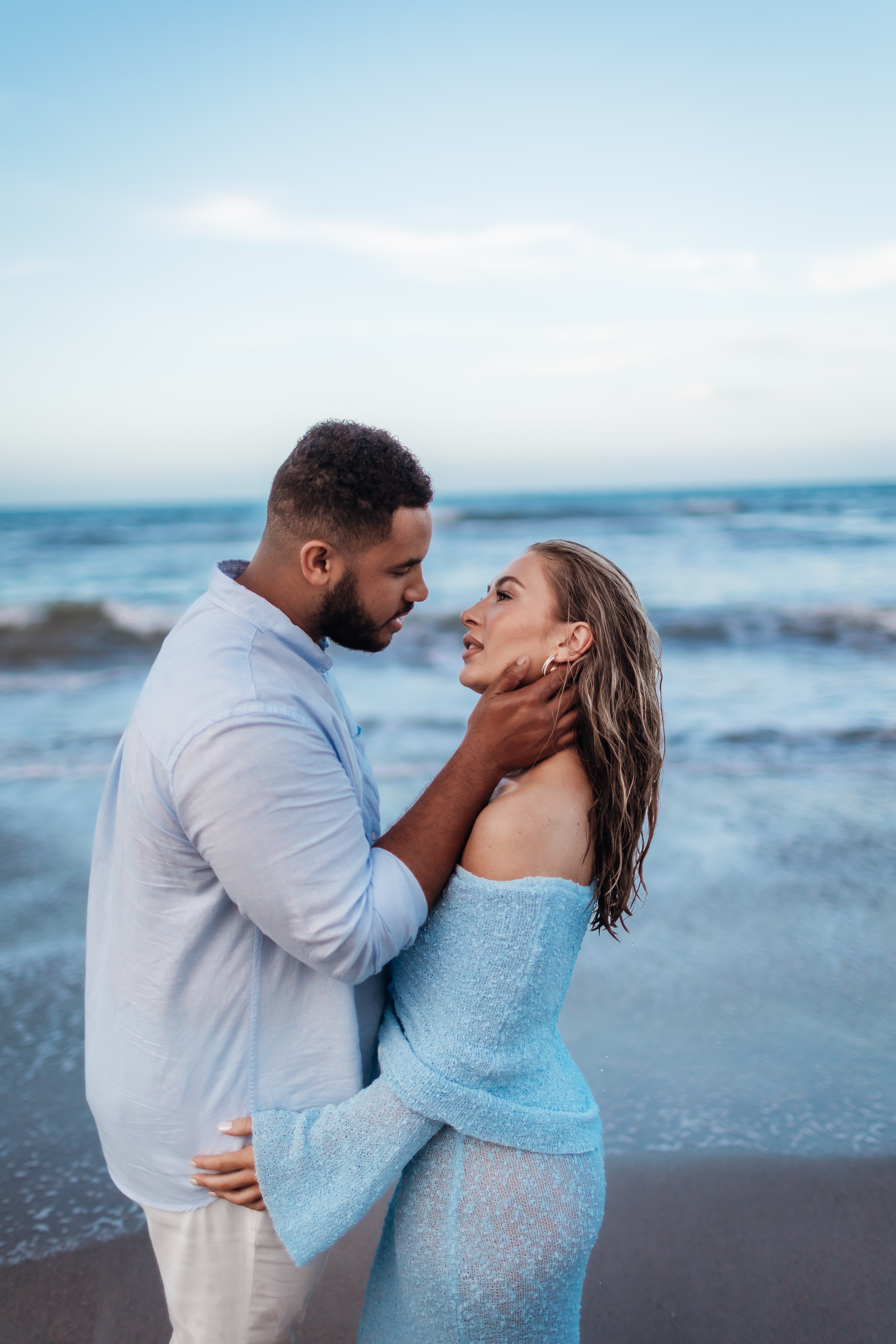 A couple stands close on the beach, facing each other, with the ocean and sky in the background.