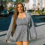 Smiling woman with long braids walking on a residential street, sunny day, houses and cars in background.