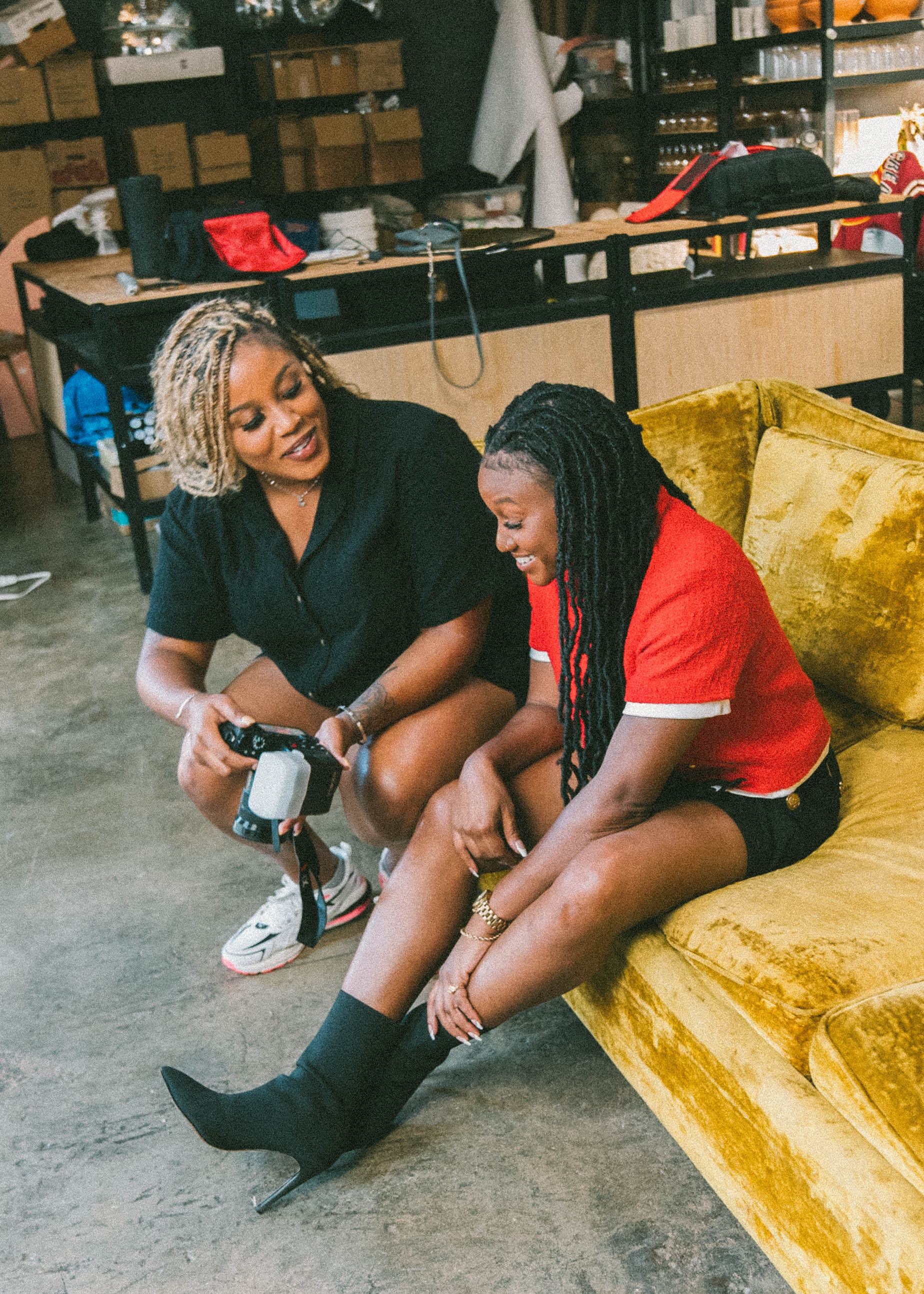 Two women sitting on a yellow couch, one adjusting a camera, in a room with shelves and boxes in the background.