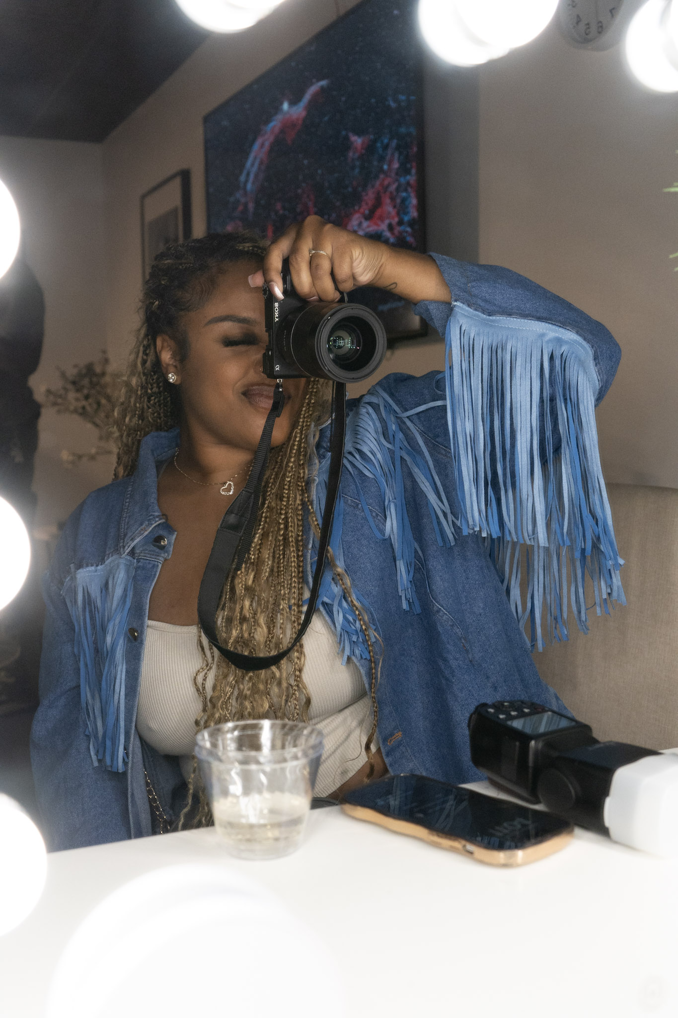 Young woman with braided hair takes a mirror selfie in a room, wearing a blue fringed jacket and white top.