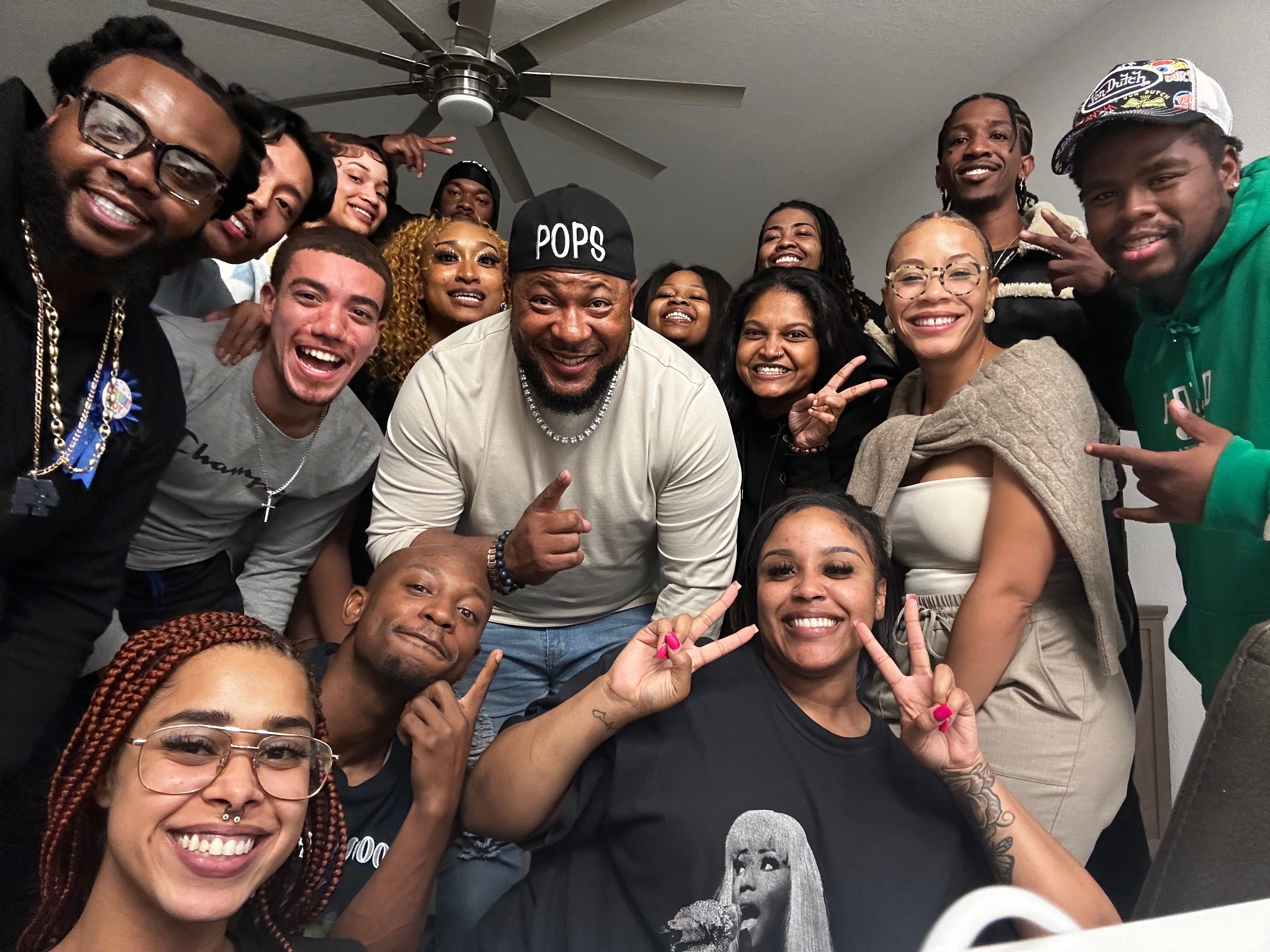 Group of people lying on the floor, smiling and making peace signs, with a ceiling fan above them.