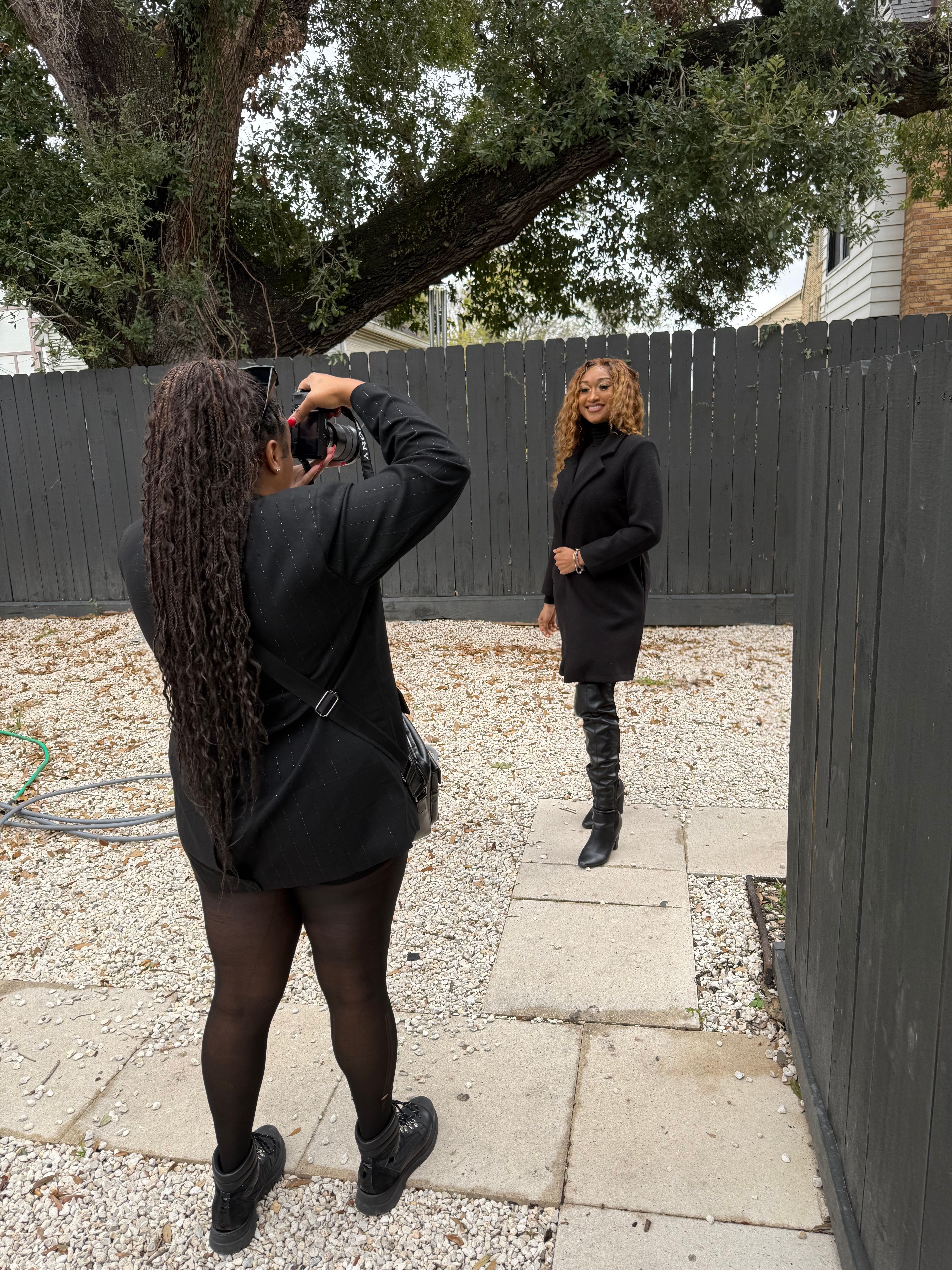 Two women standing outdoors near a black fence, one with long curly hair and the other with short hair, both dressed in black.