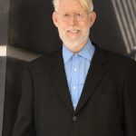 Older man with glasses, white hair, and beard, wearing a dark suit and blue shirt, smiling.