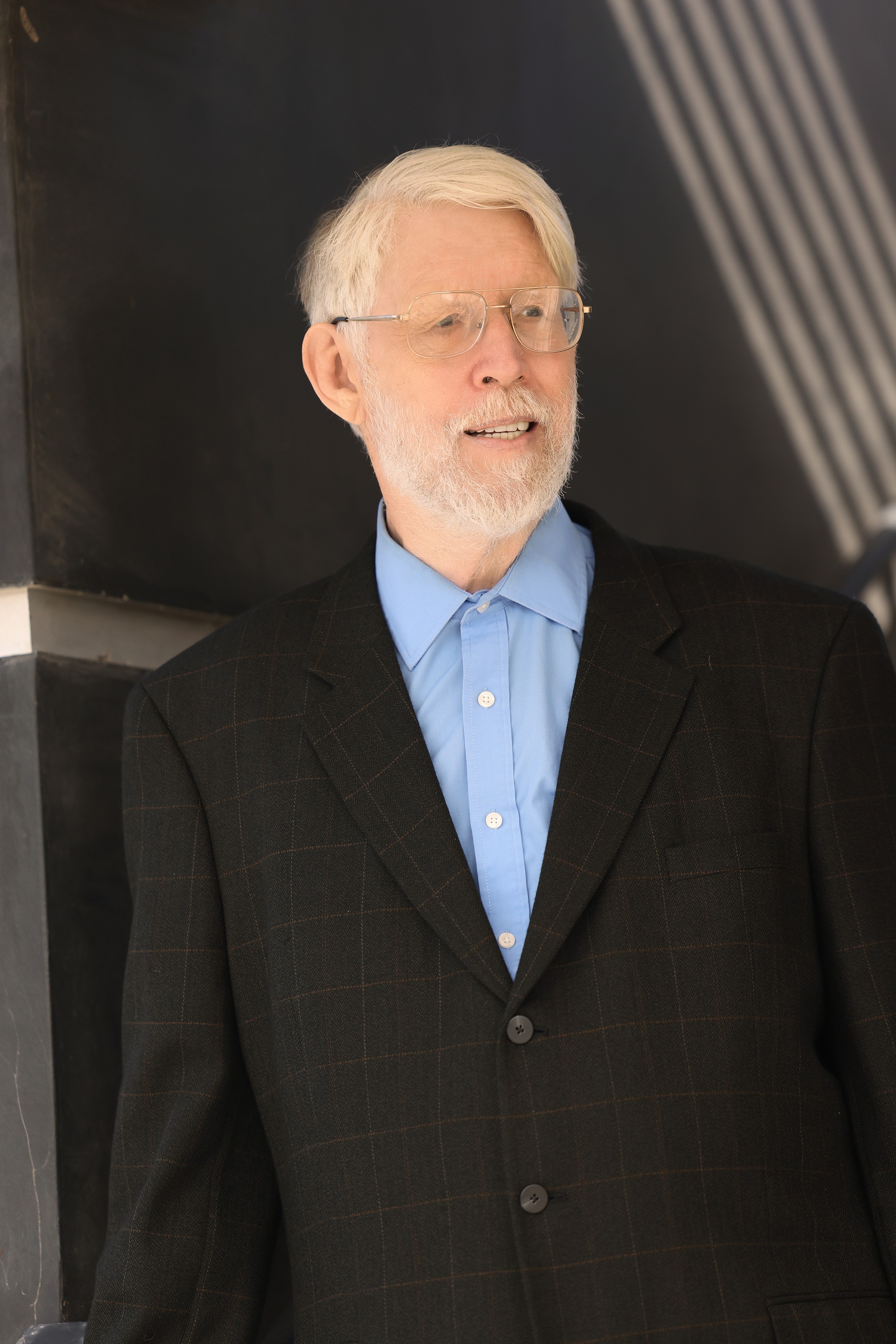 Older man with white hair and glasses wearing a dark suit and blue shirt, standing outdoors.