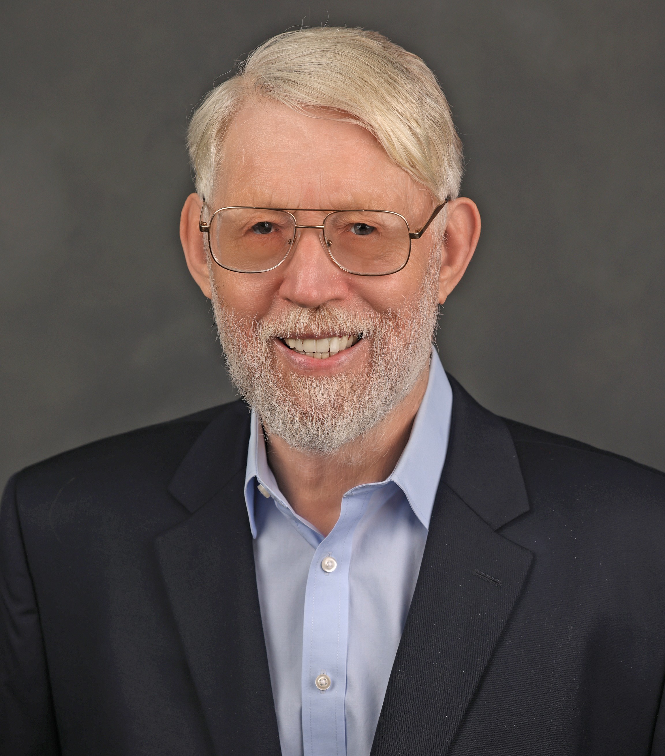 Portrait of an older man with glasses, white hair, and a beard, wearing a dark suit and light blue shirt, smiling.