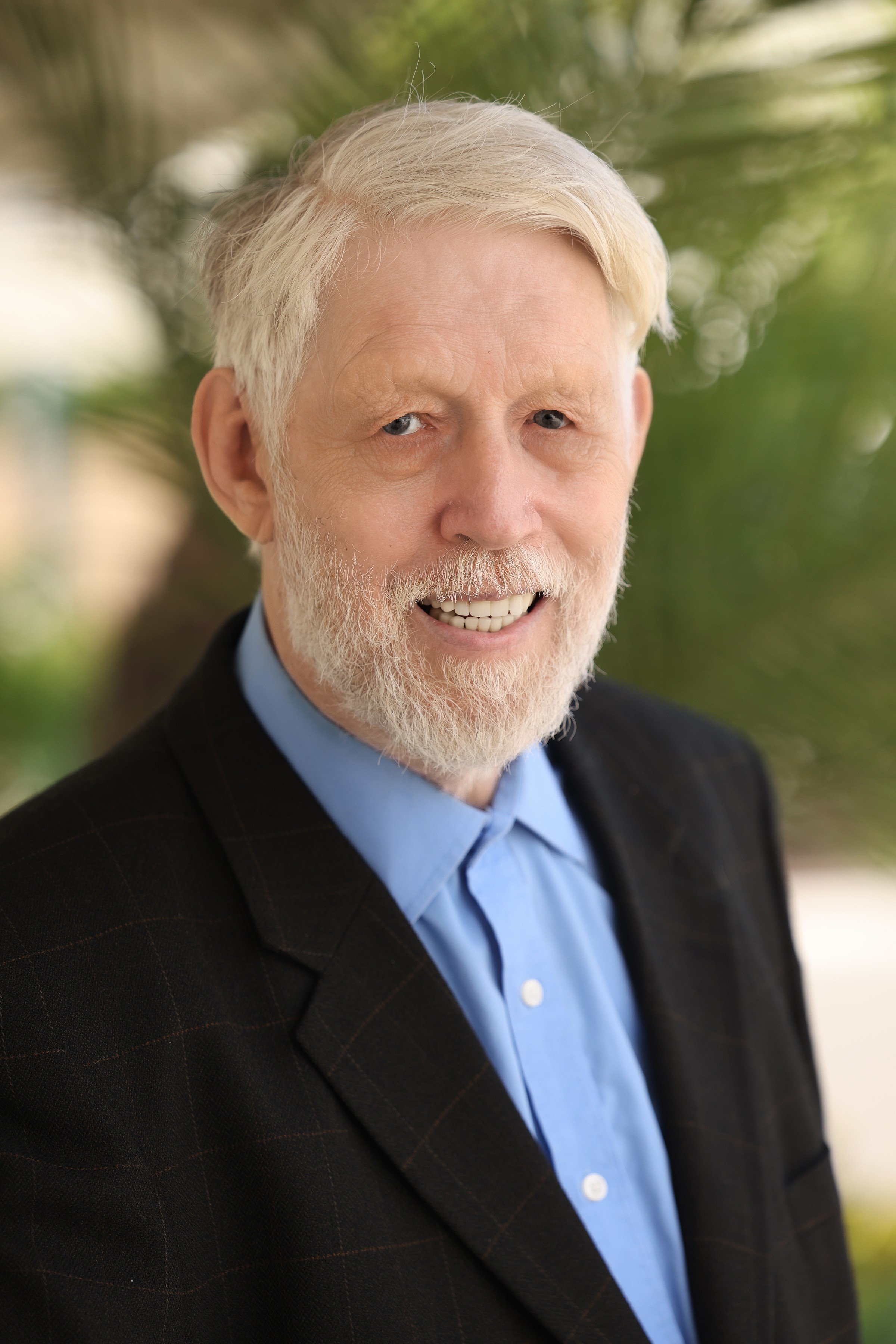 Older man with white hair and beard, smiling, wearing a black suit and blue shirt, outdoors with greenery in background.