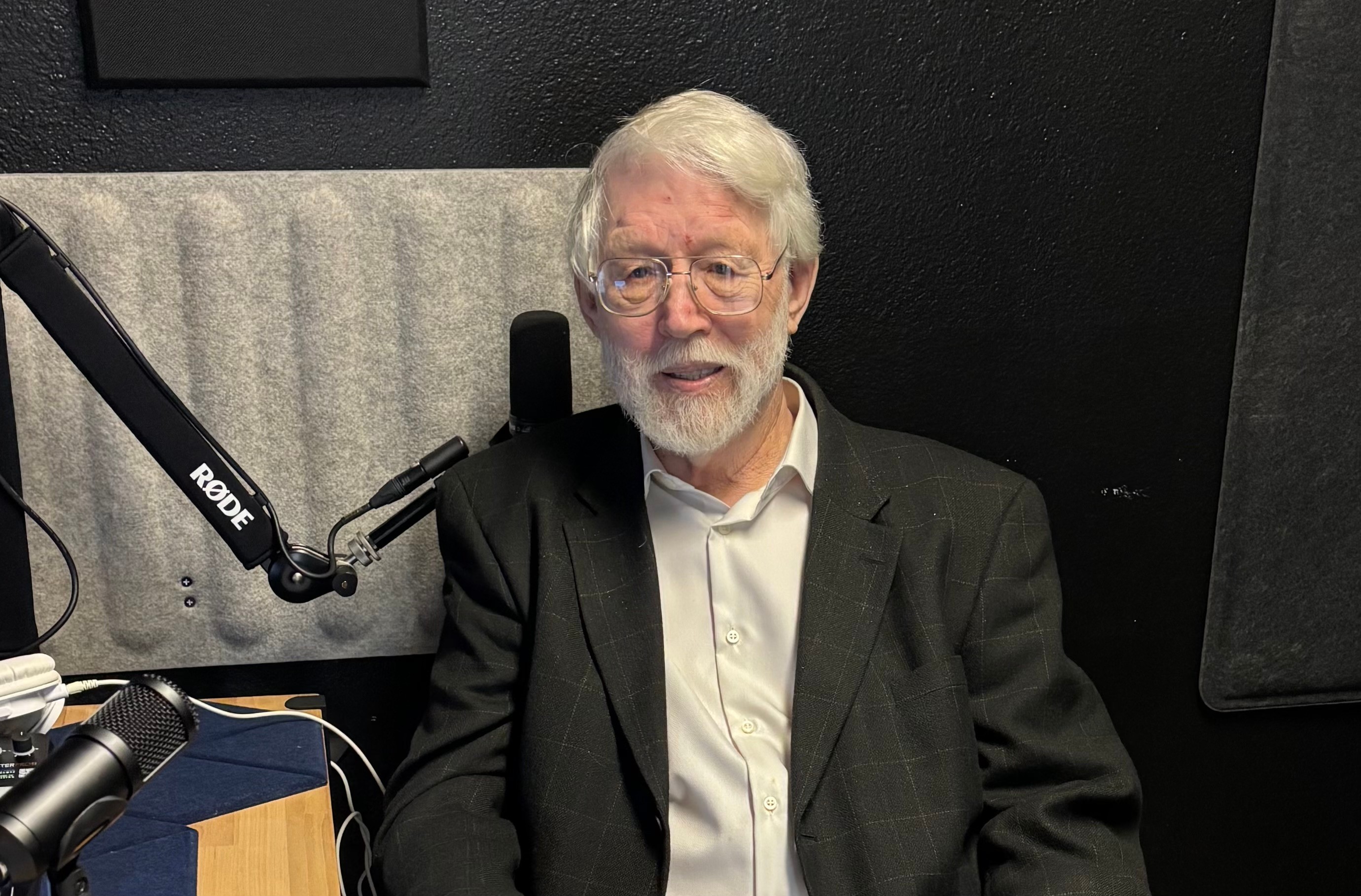 Older man with white hair and beard, wearing glasses, a white shirt, and a dark blazer, sitting in a recording studio.