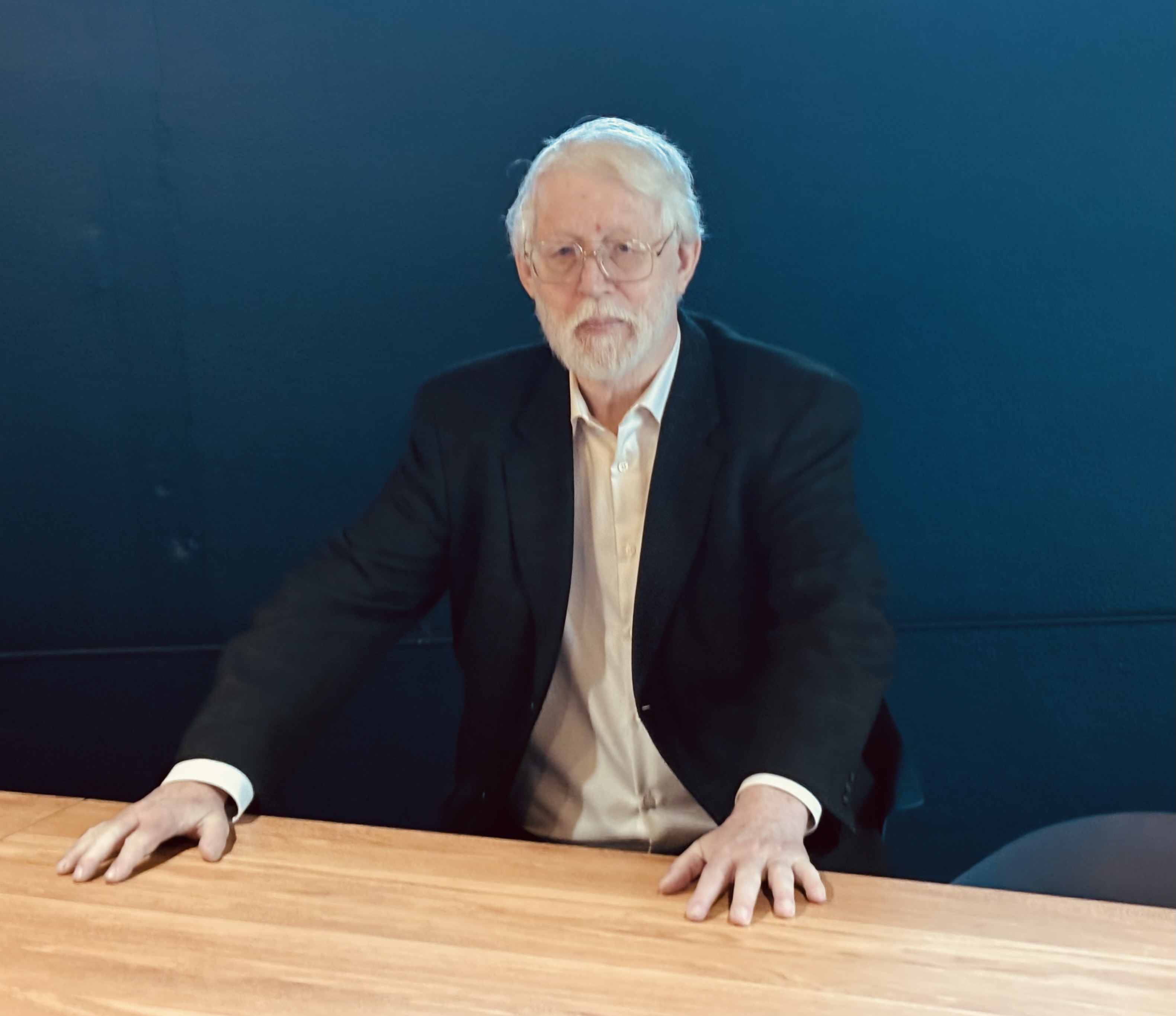 Older man with white hair and beard, wearing glasses, a dark blazer, and a white shirt, sitting at a wooden table against a dark blue background.