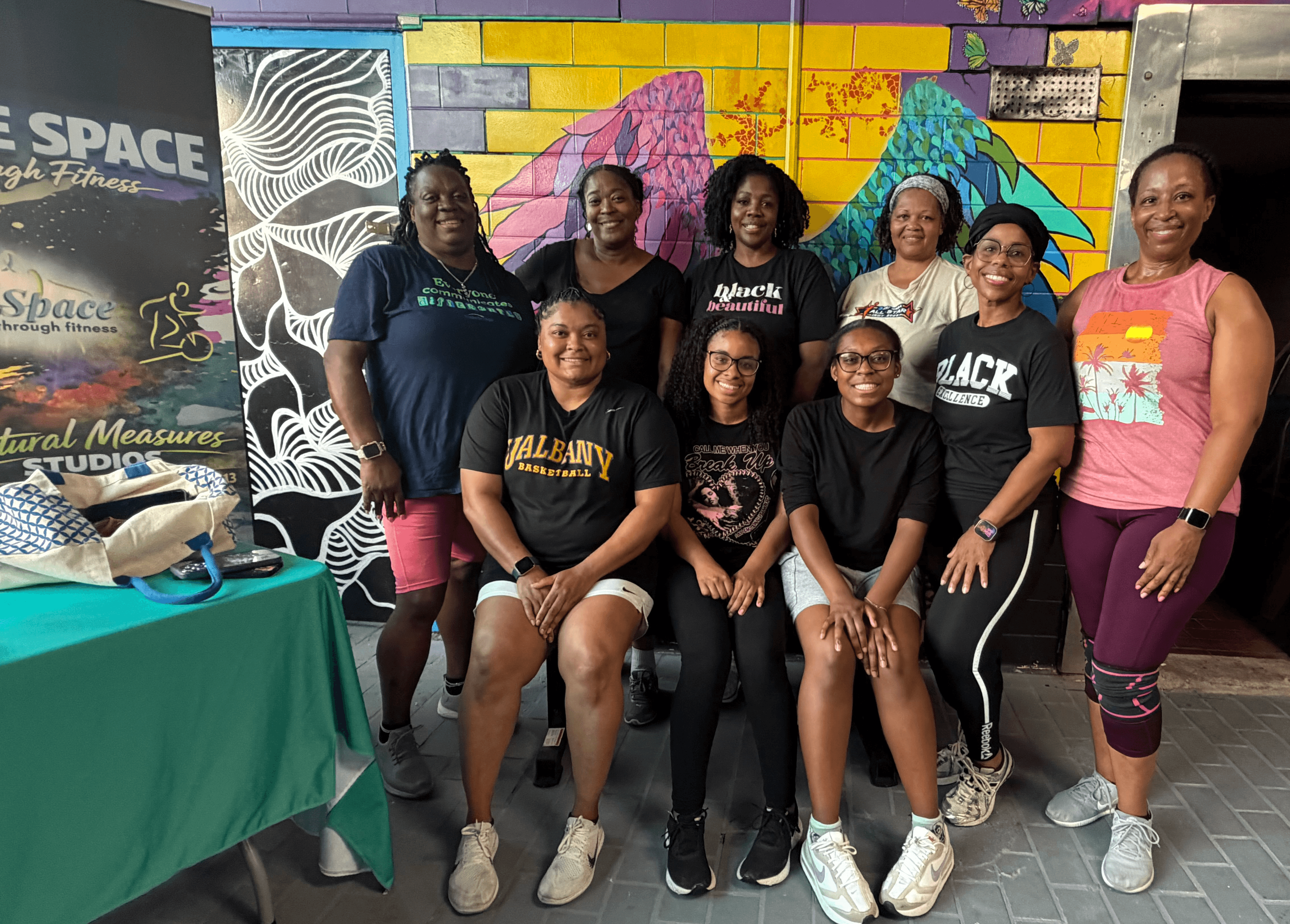 Group of nine women and girls standing and sitting in front of a colorful mural, smiling for the photo.