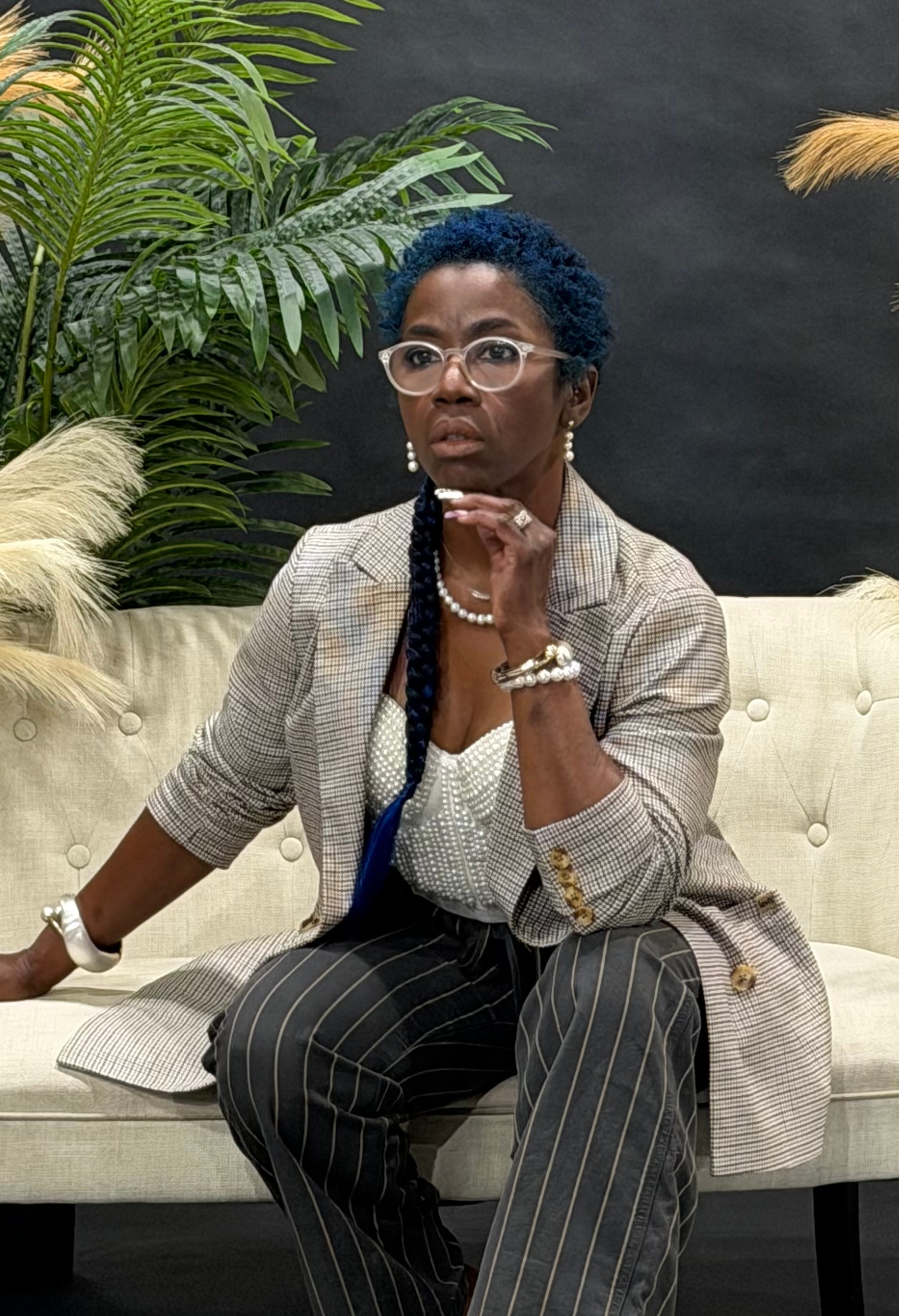 Woman with glasses and curly hair sitting on a cream-colored sofa, wearing a striped suit and pearl necklace, with plants in background.