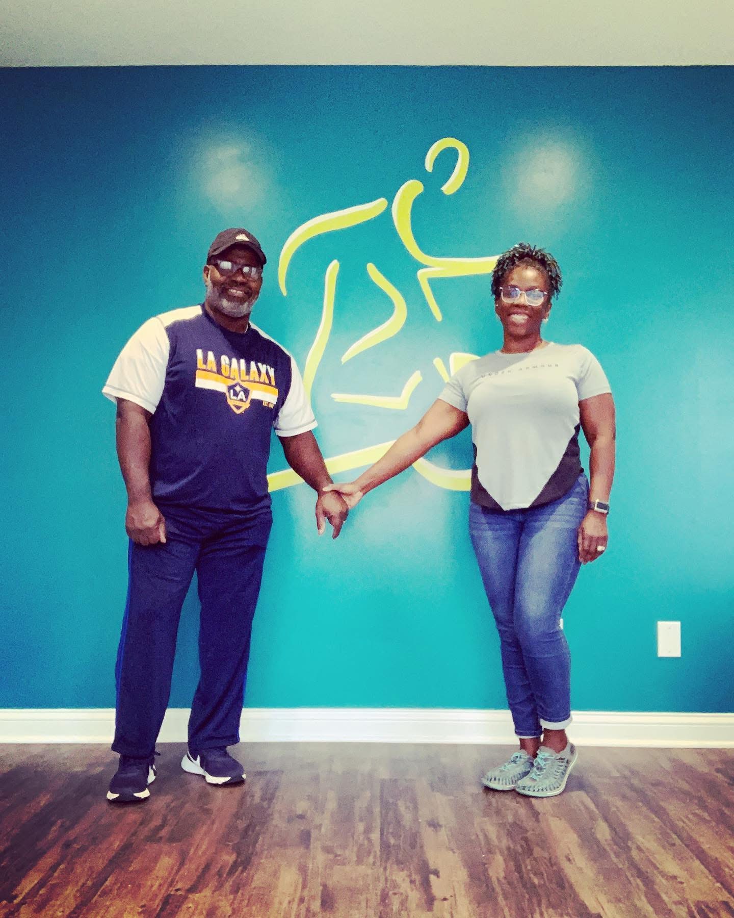 Two people holding hands in front of a blue wall with a yellow abstract figure, smiling, standing on wooden floor.