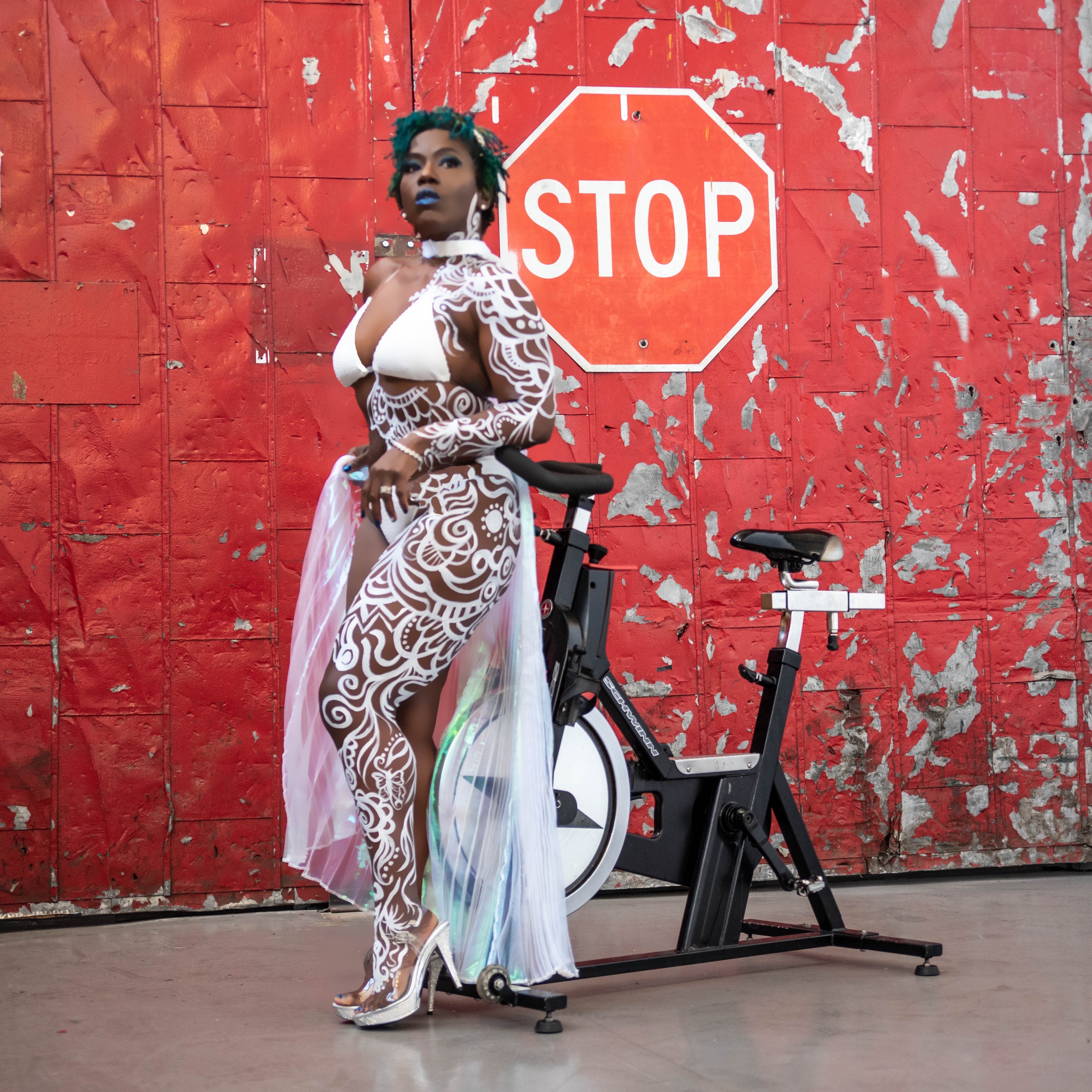 Woman in white lace outfit standing next to a stationary bike in front of a red, weathered wall with a stop sign.