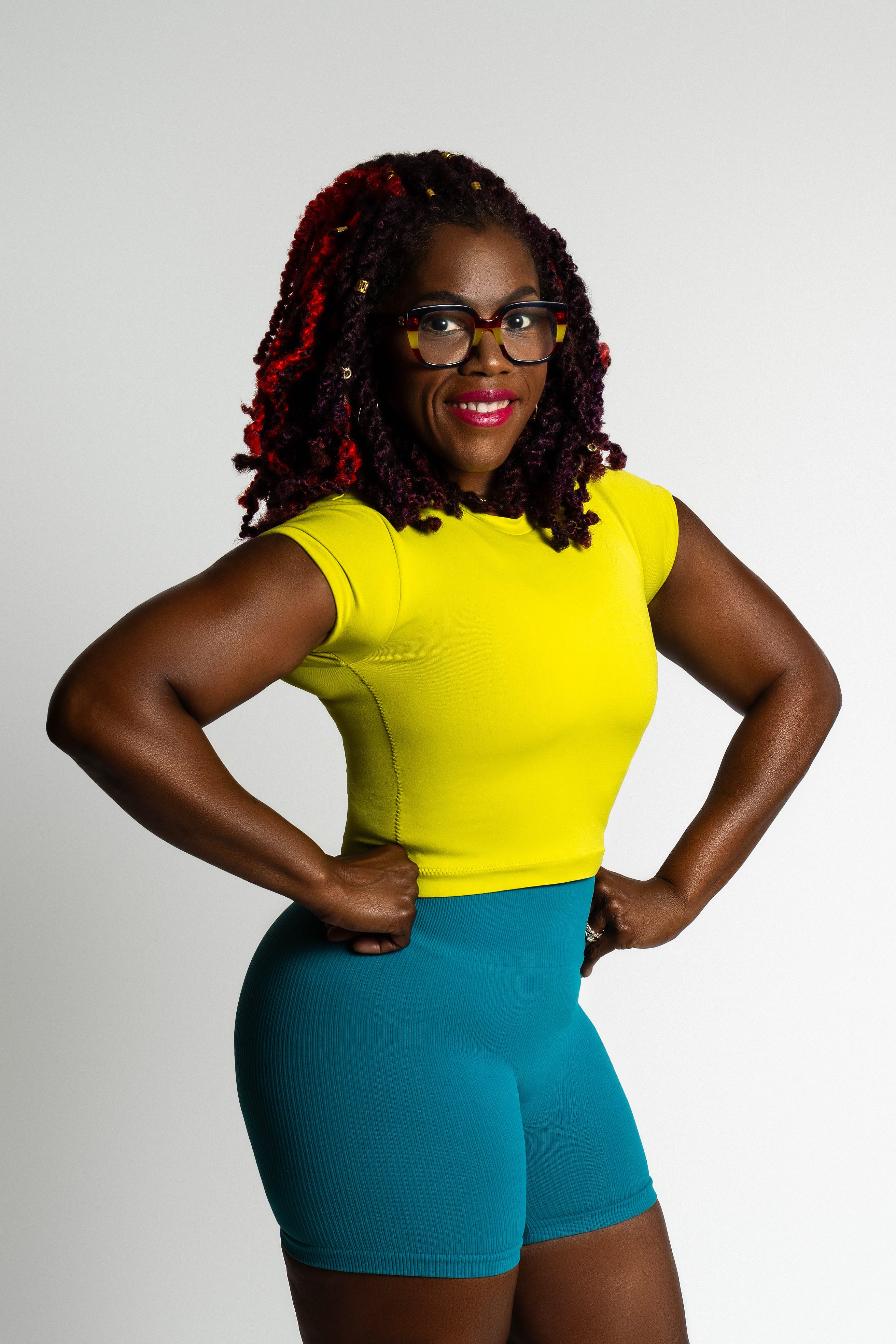 Woman with glasses and curly hair in a yellow top and teal shorts, smiling with hands on hips.