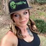 Young woman with wavy hair wearing a black cap with green skull and text, outdoors with plants and trees.