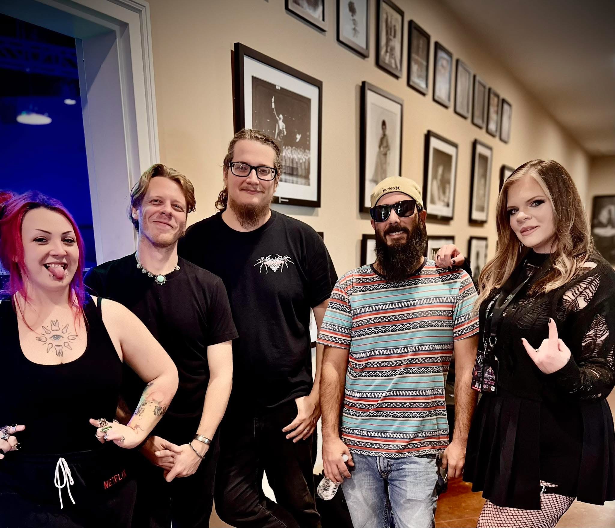 Group of five people standing indoors, smiling, with framed pictures on the wall behind them.