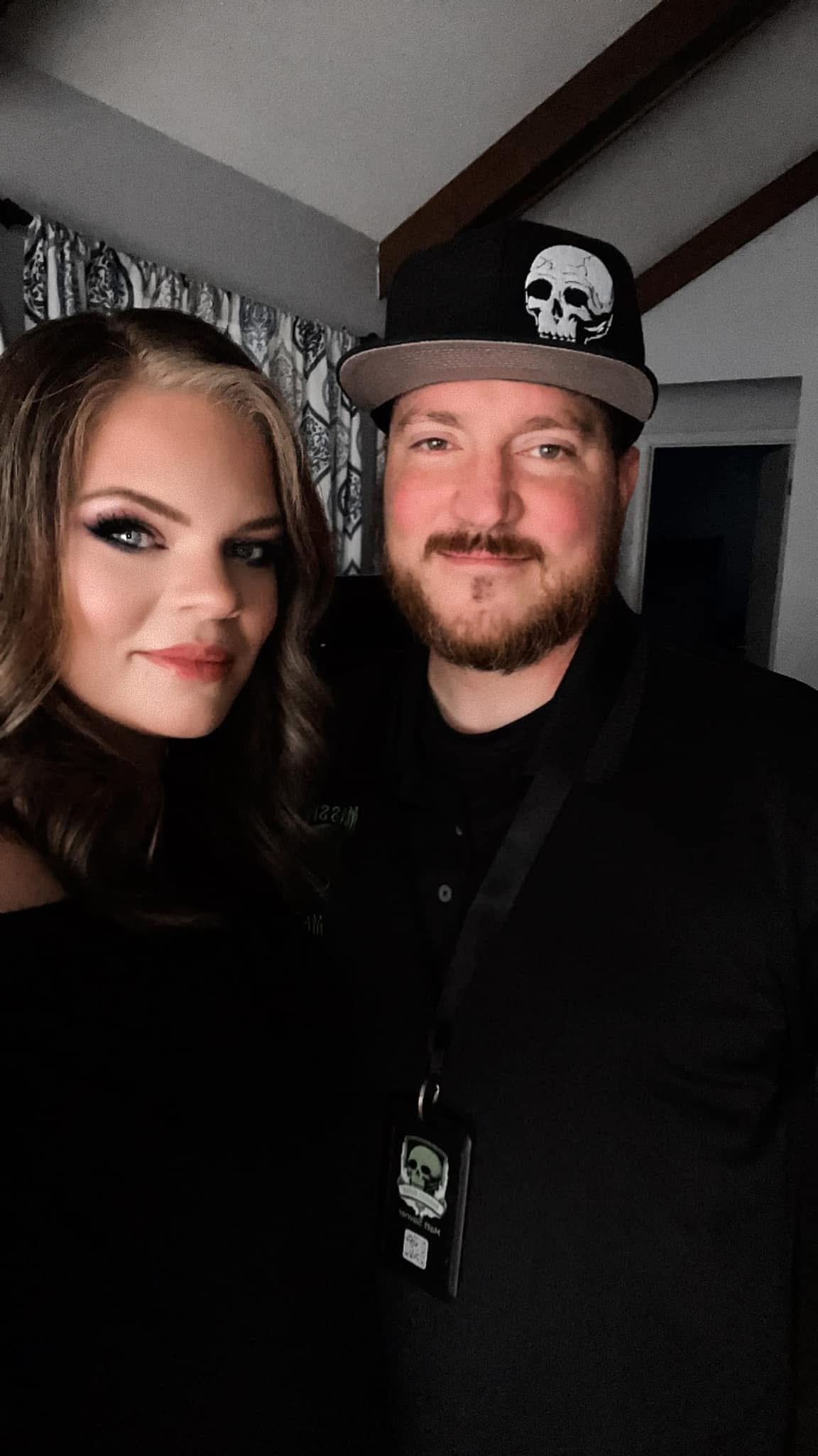 Two people posing indoors, one wearing a cap with a skull, both smiling, with a curtain and ceiling visible behind them.
