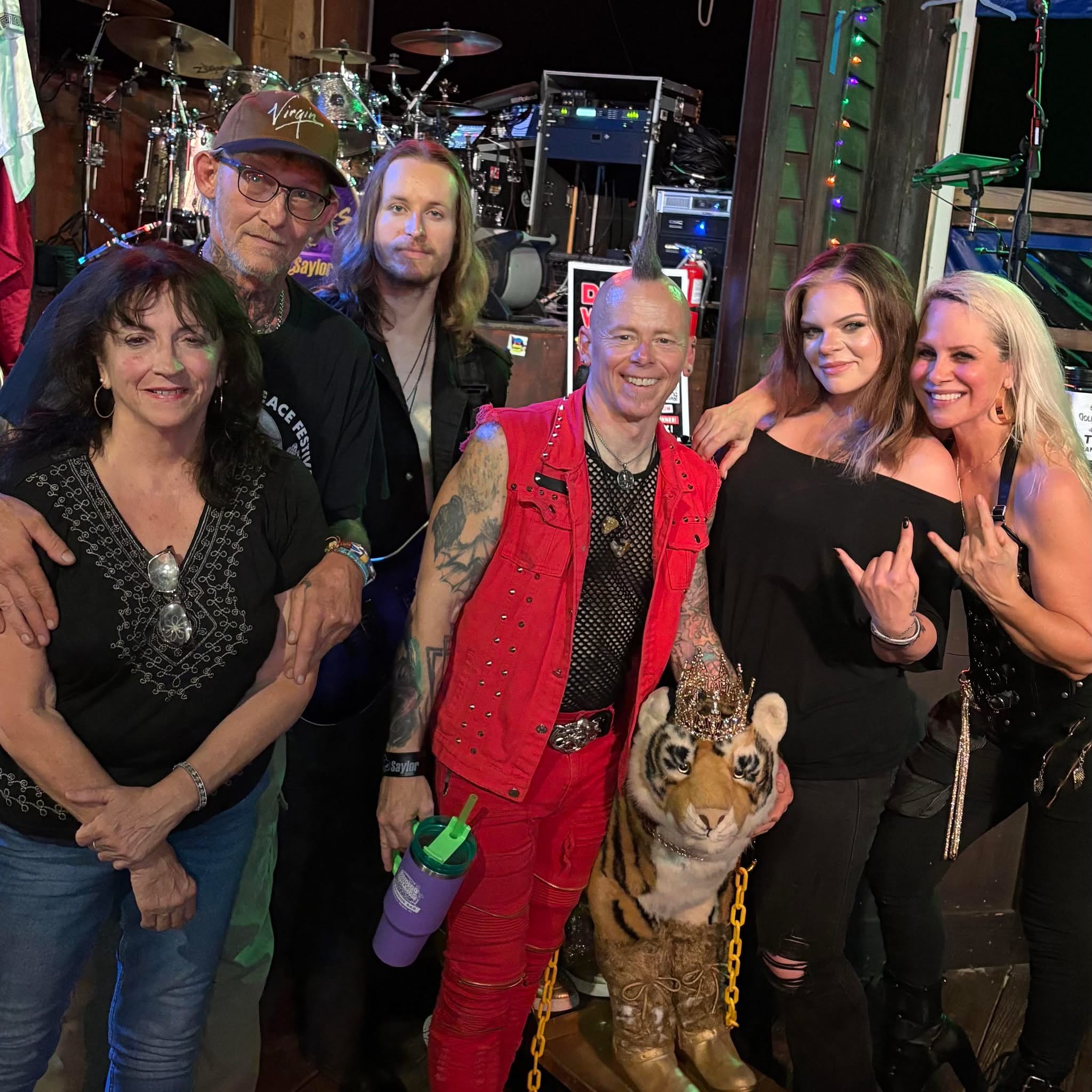 Group of six people, including a person with a dog costume, smiling and posing backstage with musical equipment in the background.