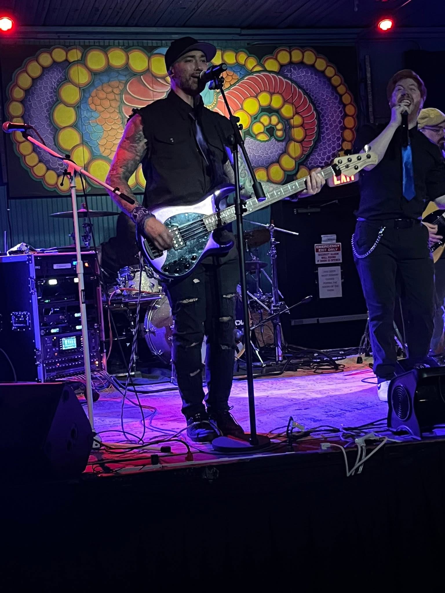Two musicians perform on stage with colorful backdrop, one playing guitar and singing, the other singing into a microphone.