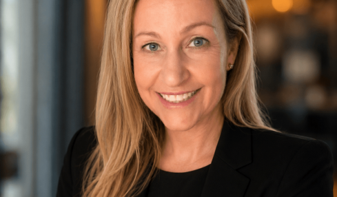 Woman with long blonde hair smiling, wearing a black blazer, in a warmly lit indoor setting.