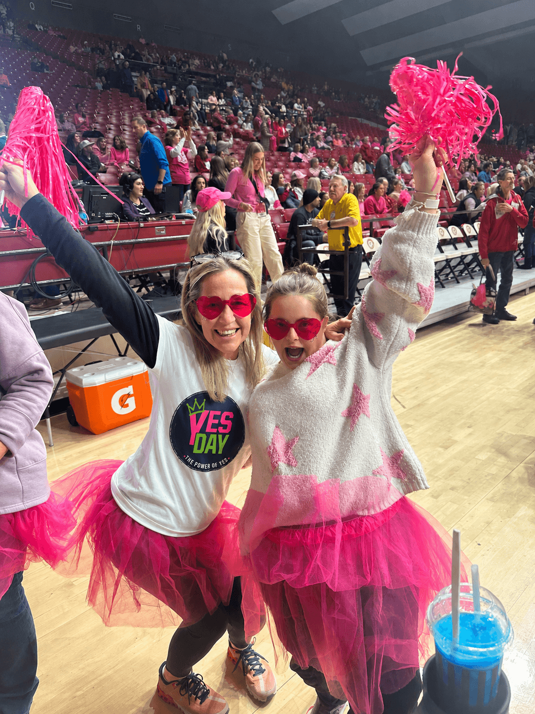 Two women in pink tutus and sweaters, wearing sunglasses, holding pink pom-poms, smiling at a sports event with a crowd in the background.