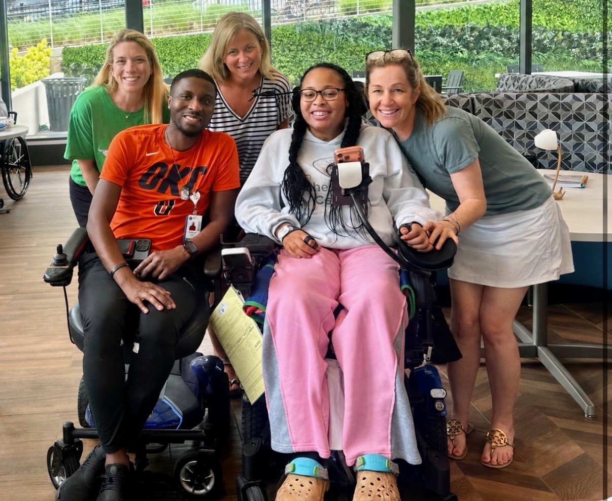 Group of five women, one in a wheelchair, smiling indoors with large windows and greenery outside, casual clothing.