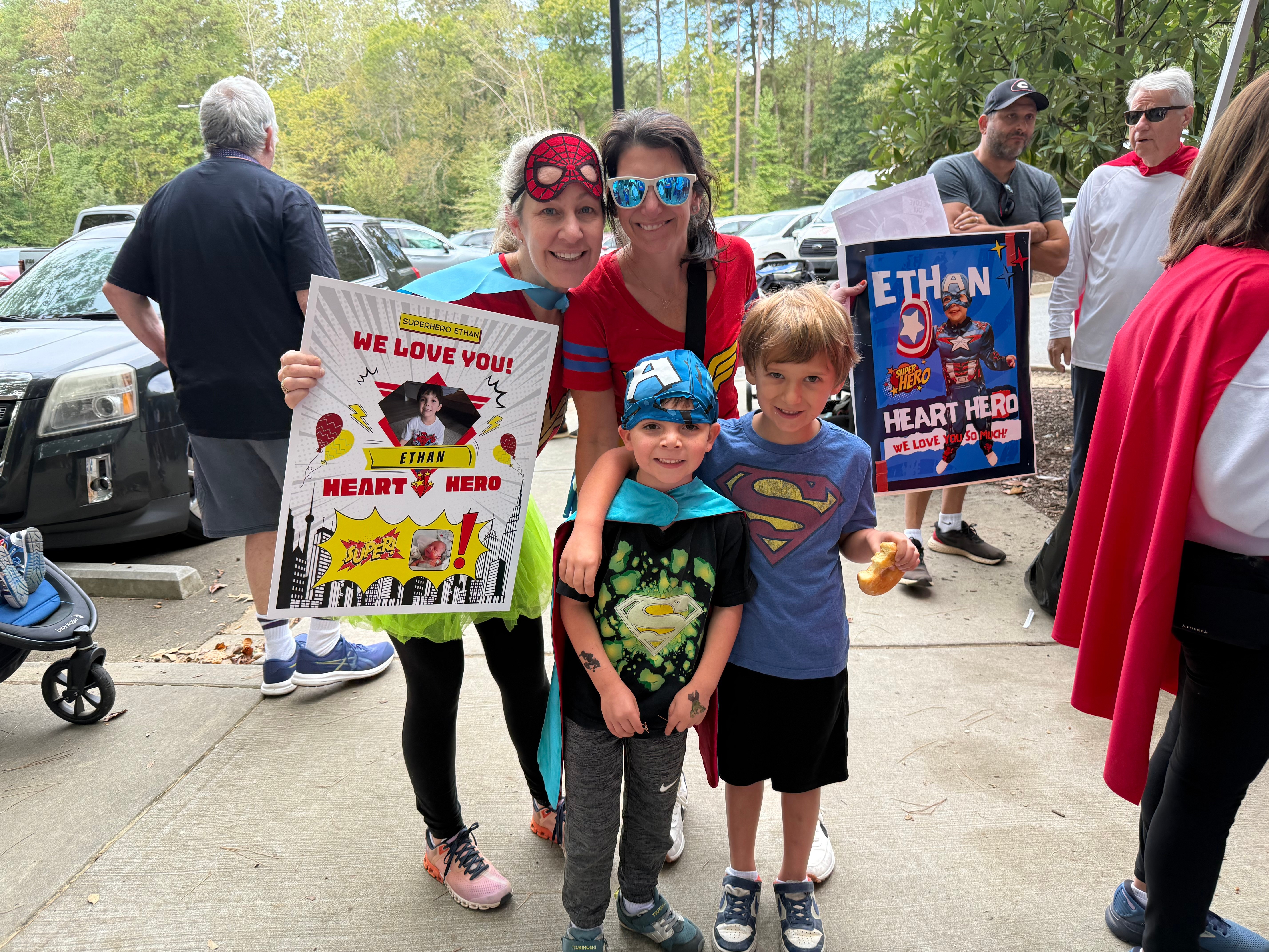 Children and adults gather outdoors, holding colorful superhero-themed signs, with trees and parked cars in the background.
