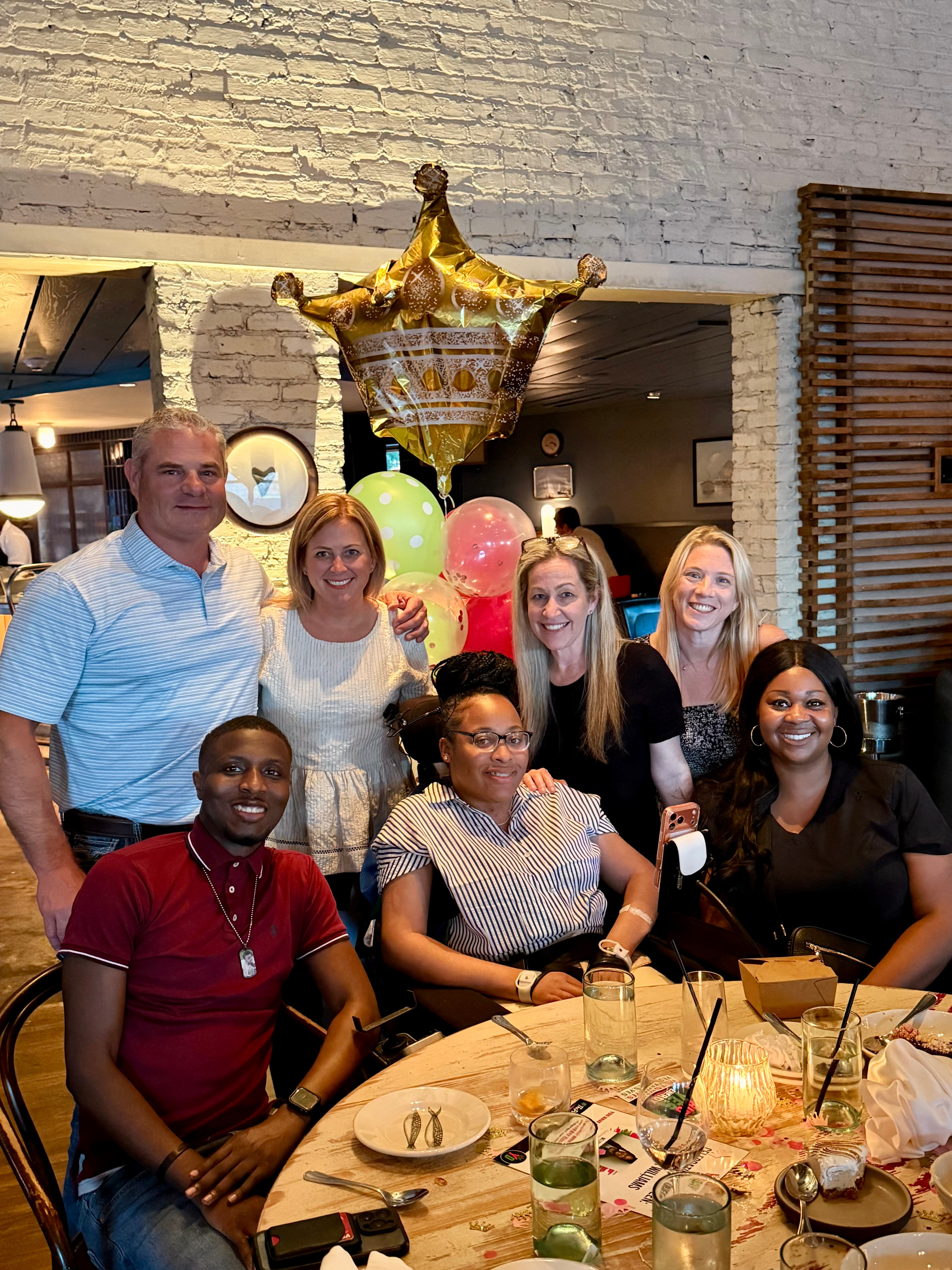 Group of seven people gathered around a table with drinks and food, celebrating with balloons and a gold balloon decoration, in a decorated indoor space.