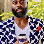 Man with a beard and bald head holding a small container outdoors, wearing a patterned blazer and white shirt, with green foliage in background.