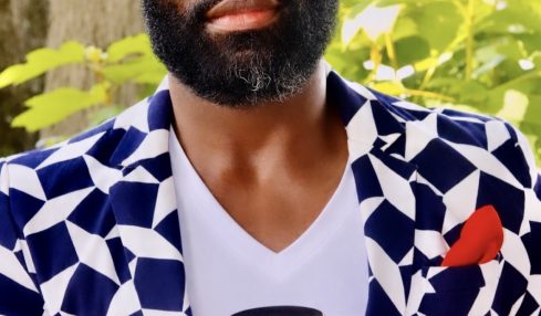 Man with a beard and bald head holding a small container outdoors, wearing a patterned blazer and white shirt, with green foliage in background.