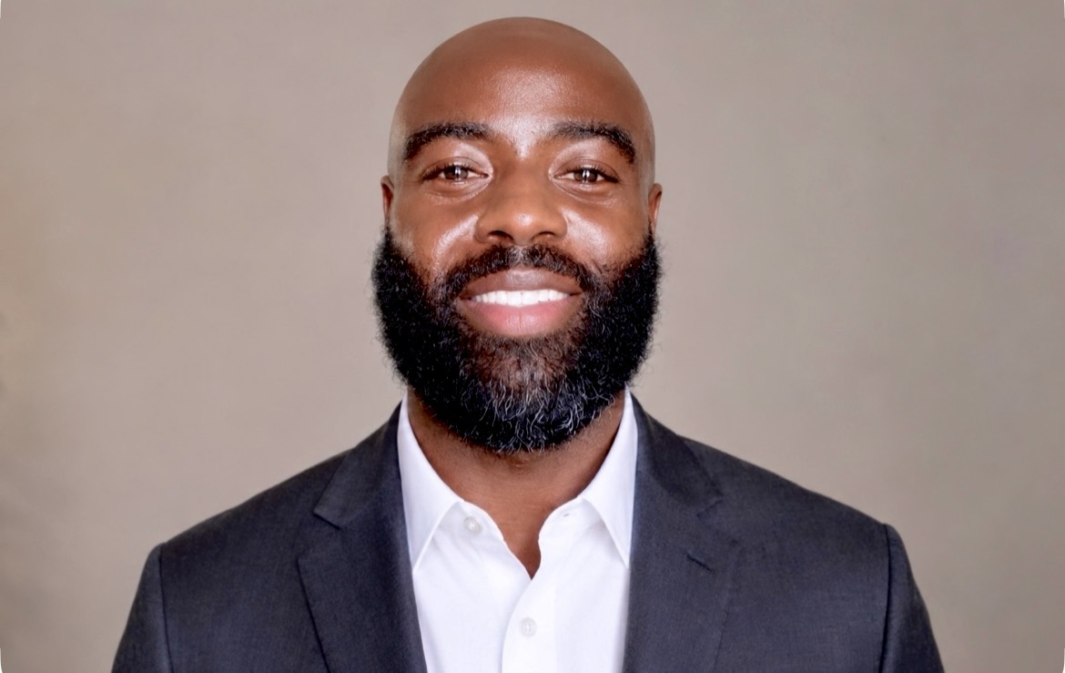 Portrait of a smiling man with a beard wearing a suit and white shirt against a plain background.