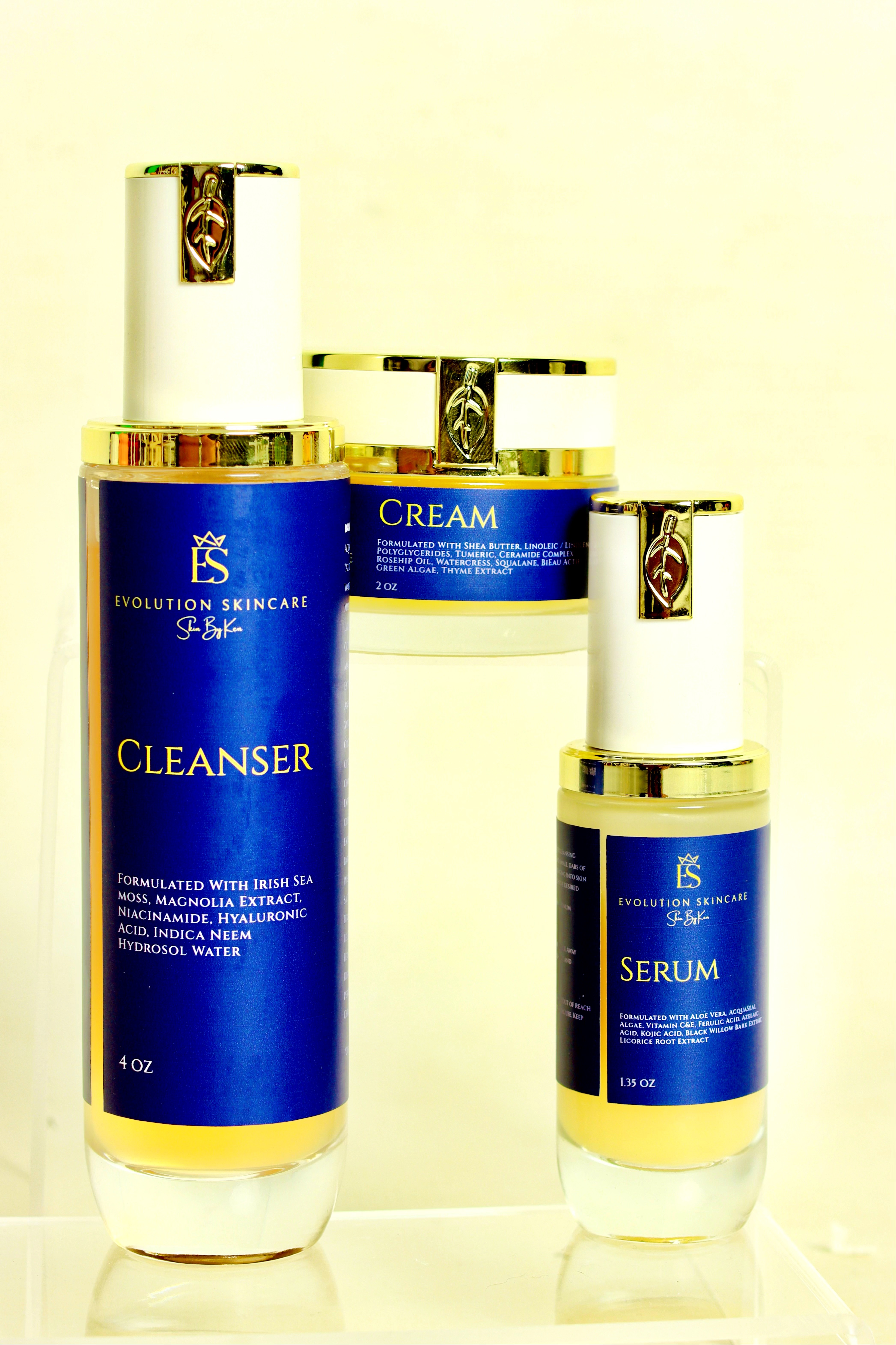 Three skincare bottles with blue labels and white caps, labeled cleanser, cream, and serum, on a light background.